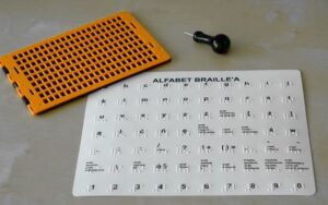 Tabliczka z przedstawionym alfabetem, dłutko i tabliczka do pisania brajlem.