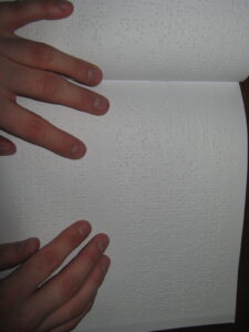 Książka brajlowska, a na niej dwie czytające dłonie.